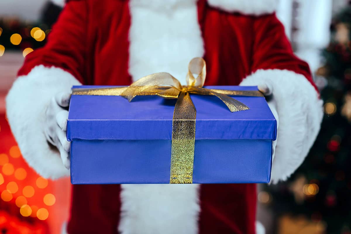 A person in a red Santa suit holds a blue gift box with a gold ribbon in front of a festive background with blurred lights.